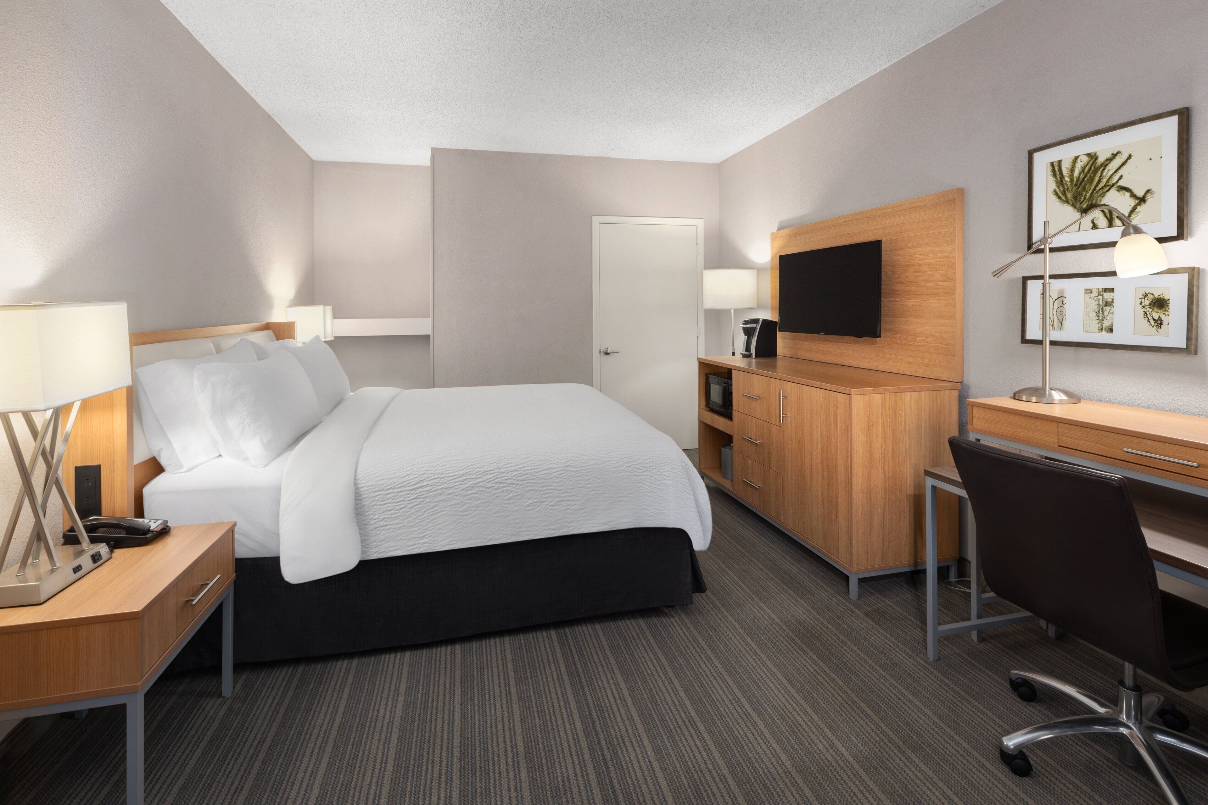 Quarto de hotel moderno com uma cama bem arrumada, uma escrivaninha com computador, televisão e um pequeno armário.