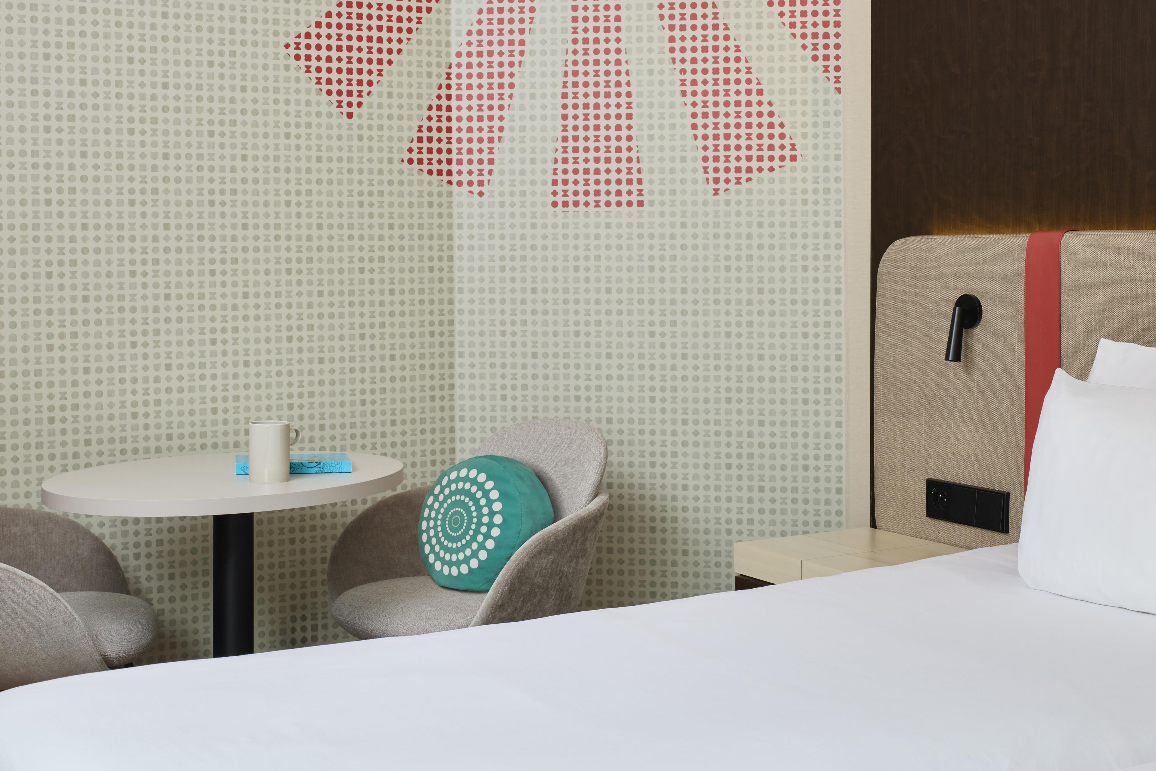A hotel room with a bed, a small table, a chair, and a wall with a geometric pattern.