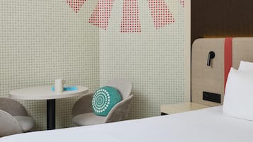 A hotel room with a bed, a small table, a chair, and a wall with a geometric pattern.