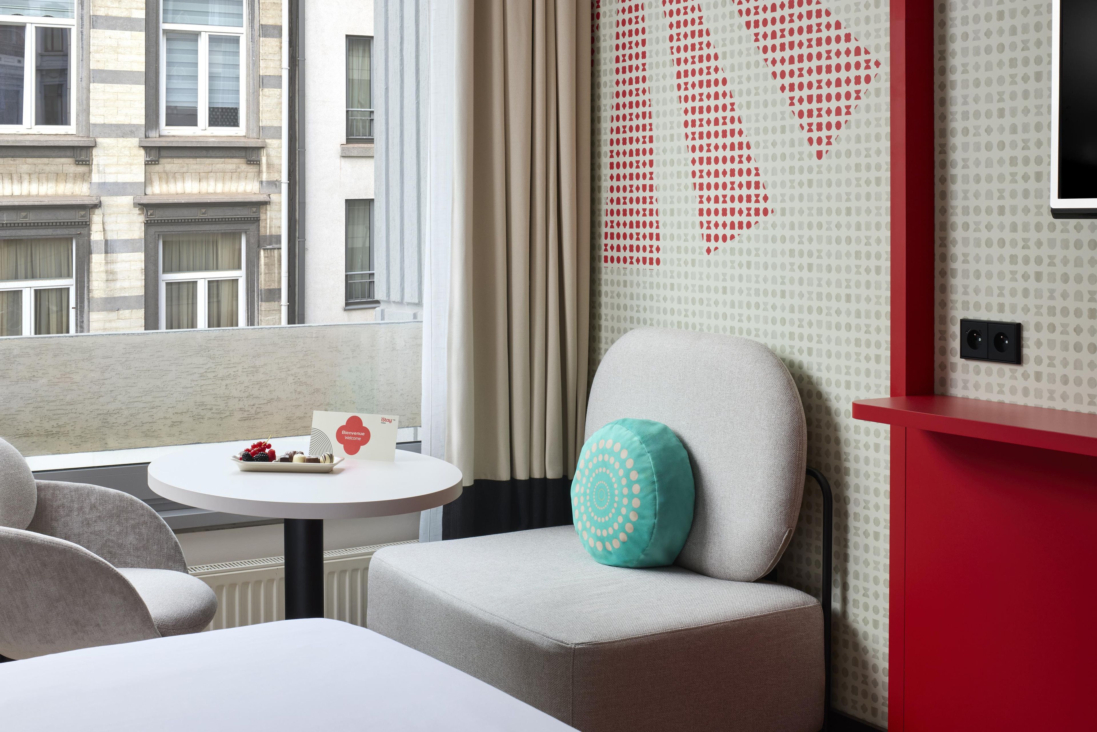 Een hotelkamer met uitzicht op de stad, een ronde tafel met een welkomstkaart, een blauwgroen kussen en een rode accentmuur.