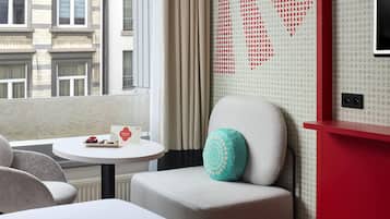 Een hotelkamer met uitzicht op de stad, een ronde tafel met een welkomstkaart, een blauwgroen kussen en een rode accentmuur.