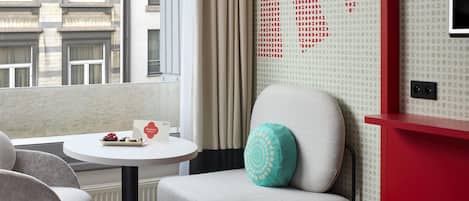 A hotel room with a view of the city, a round table with a welcome card, a teal pillow, and a red accent wall.