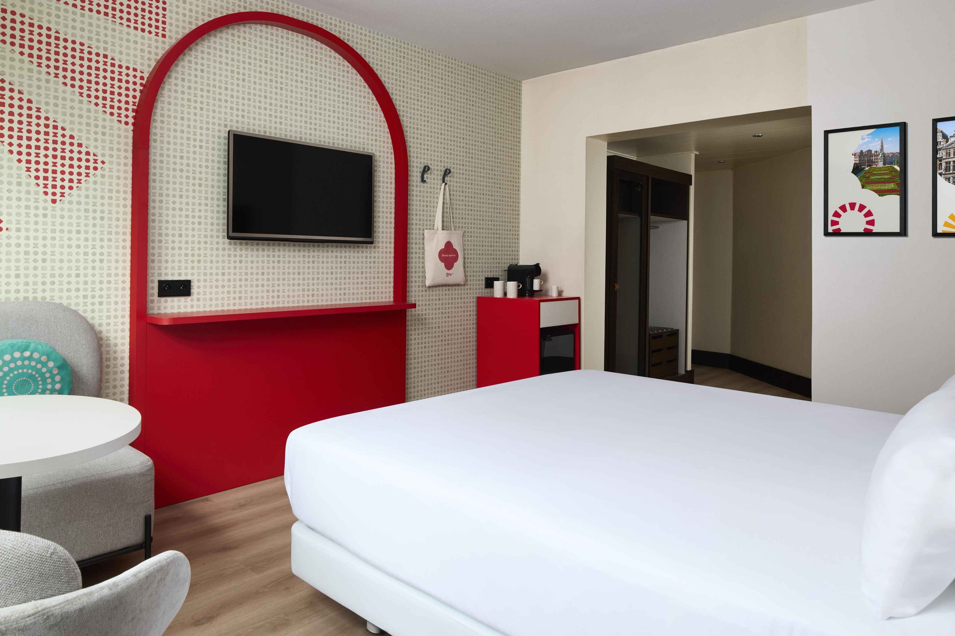 Ein Hotelzimmer mit einem Bett, einem an einer roten Wand montierten Fernseher, einem kleinen Schreibtisch mit Kaffeemaschine und einer Tür zu einem Badezimmer.
