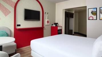 Ein Hotelzimmer mit einem Bett, einem an einer roten Wand montierten Fernseher, einem kleinen Schreibtisch mit Kaffeemaschine und einer Tür zu einem Badezimmer.