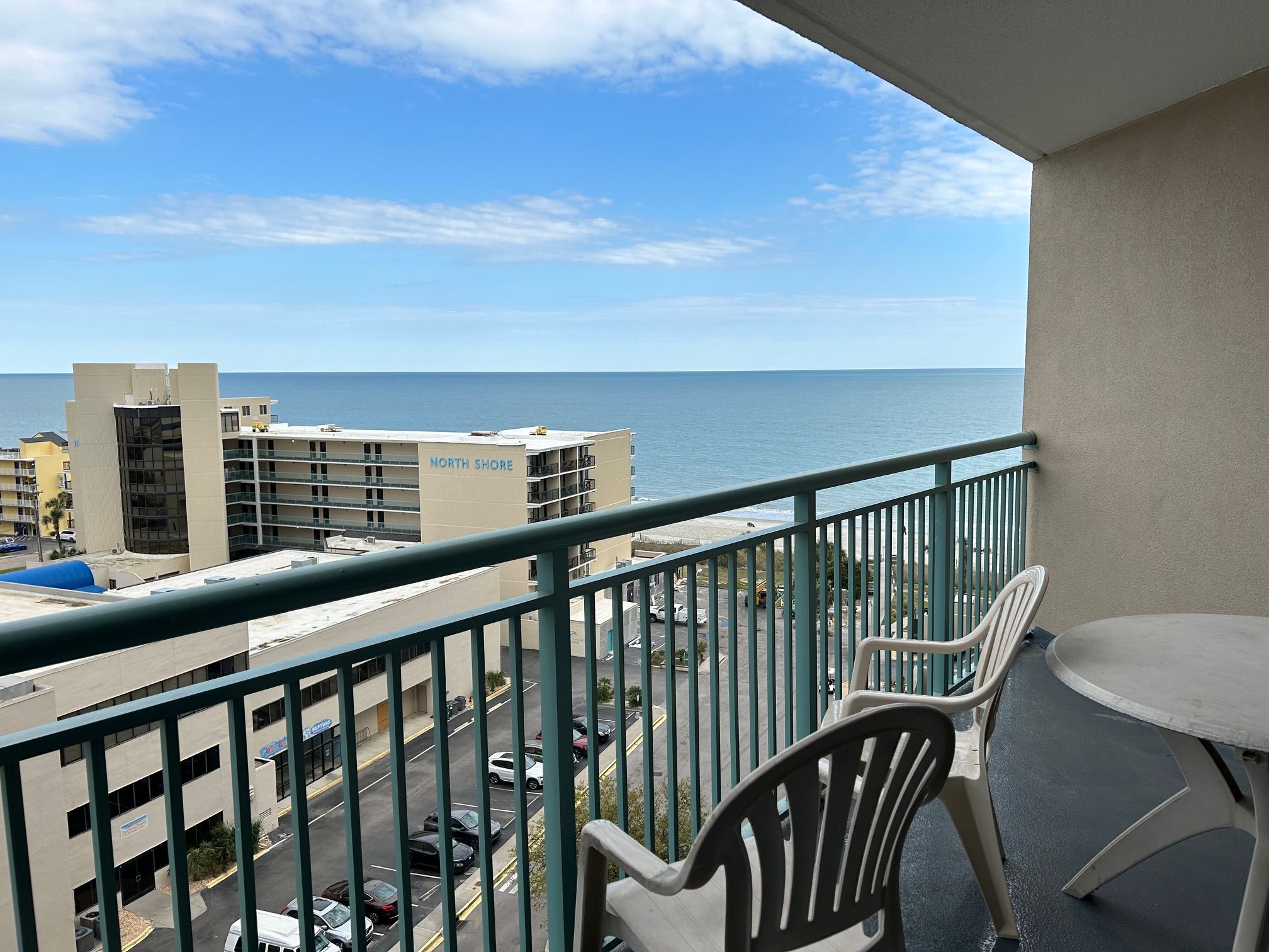 A balcony with a view of the ocean, a parking lot, and buildings in the background.
