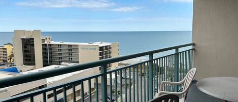 A balcony with a view of the ocean, a parking lot, and buildings in the background.