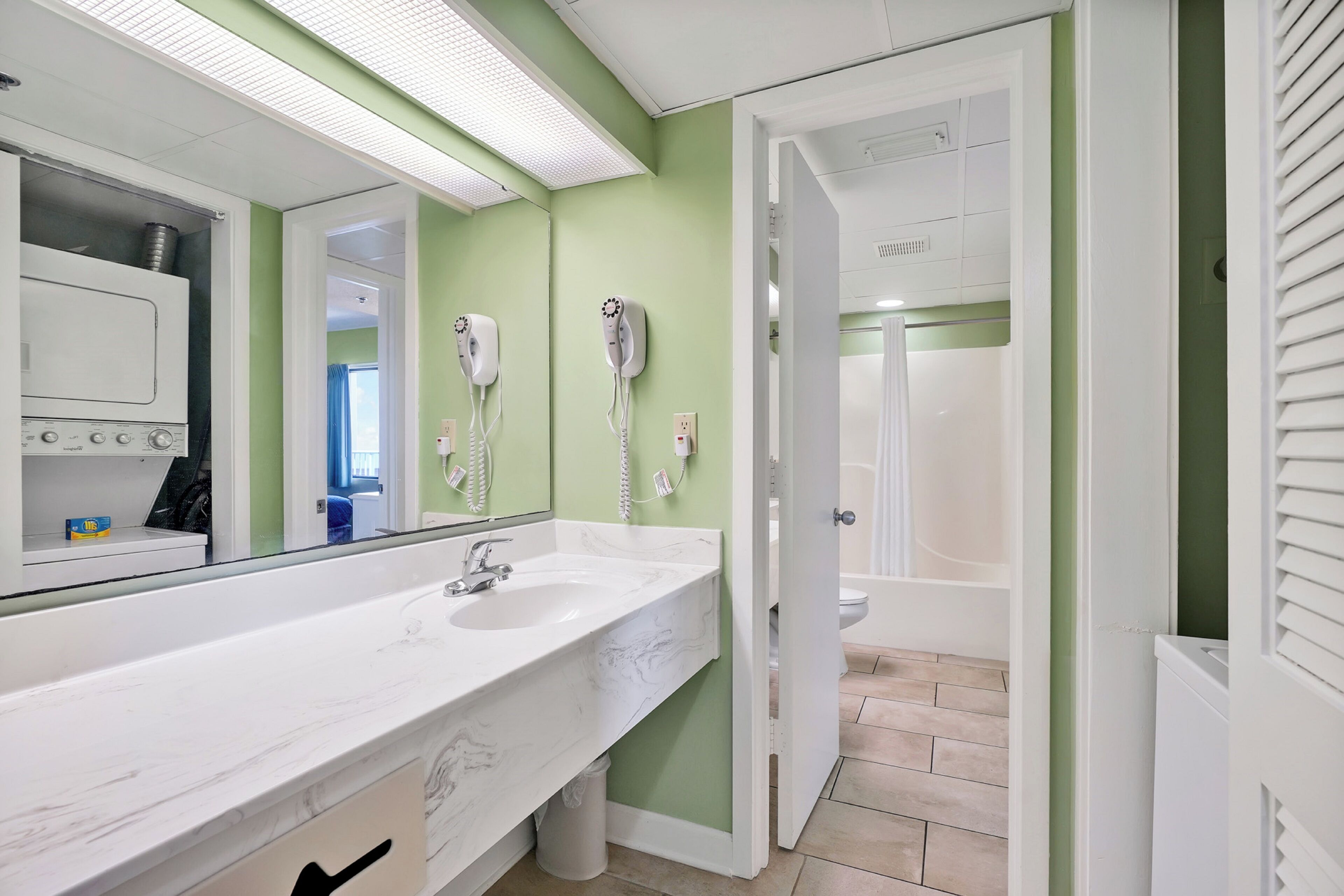 A bathroom with a white sink, a large mirror, two hair dryers mounted on the wall, and a visible shower area.
