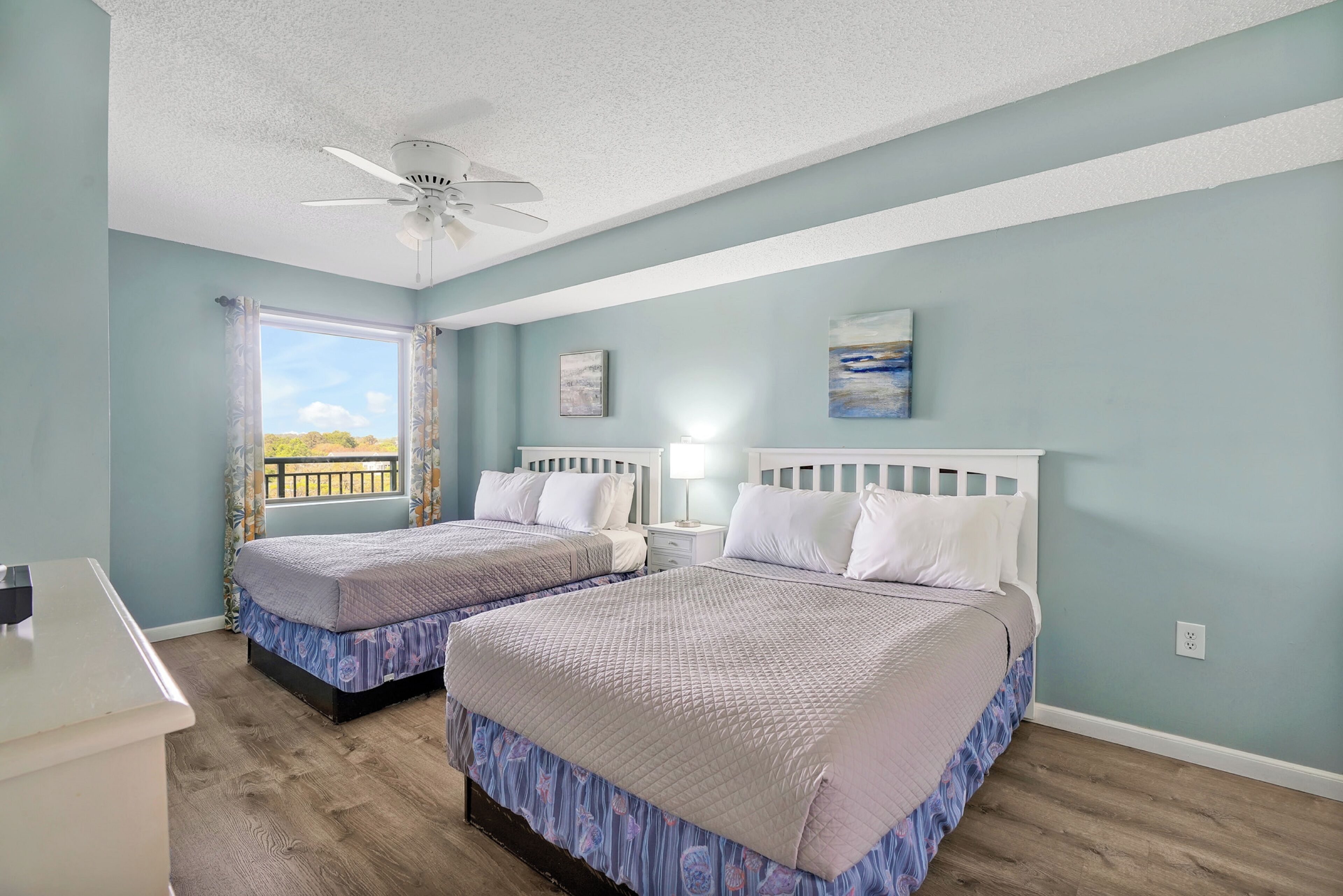 A bedroom with two beds, a ceiling fan, a window with curtains, a nightstand, and a painting on the wall.