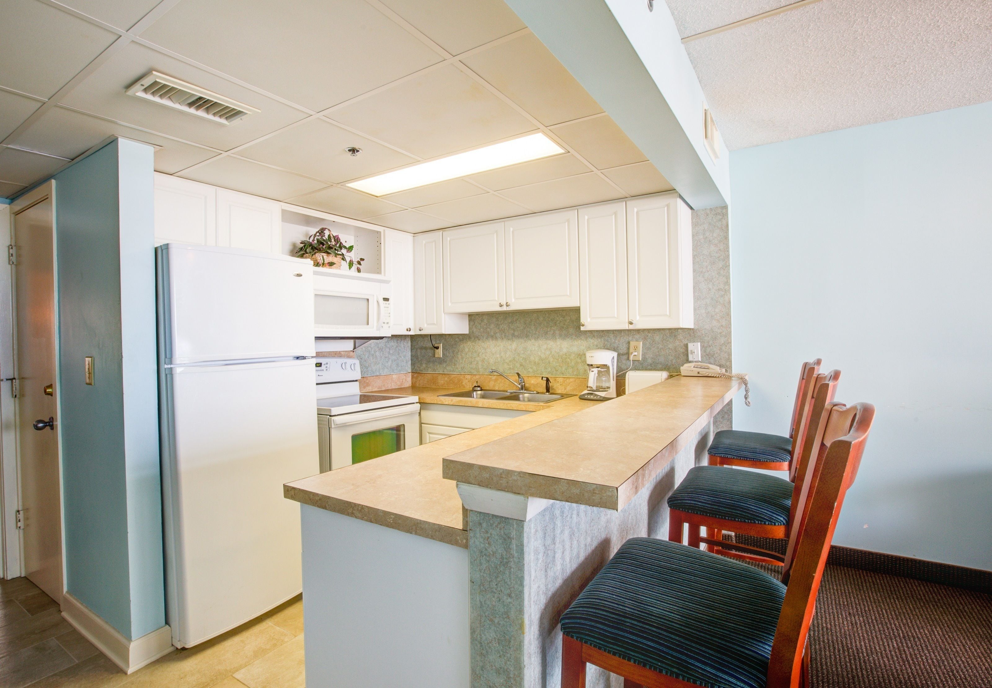 A kitchen with white cabinets, a built-in microwave, a sink, and a coffee maker. There are two wooden chairs with cushioned seats.
