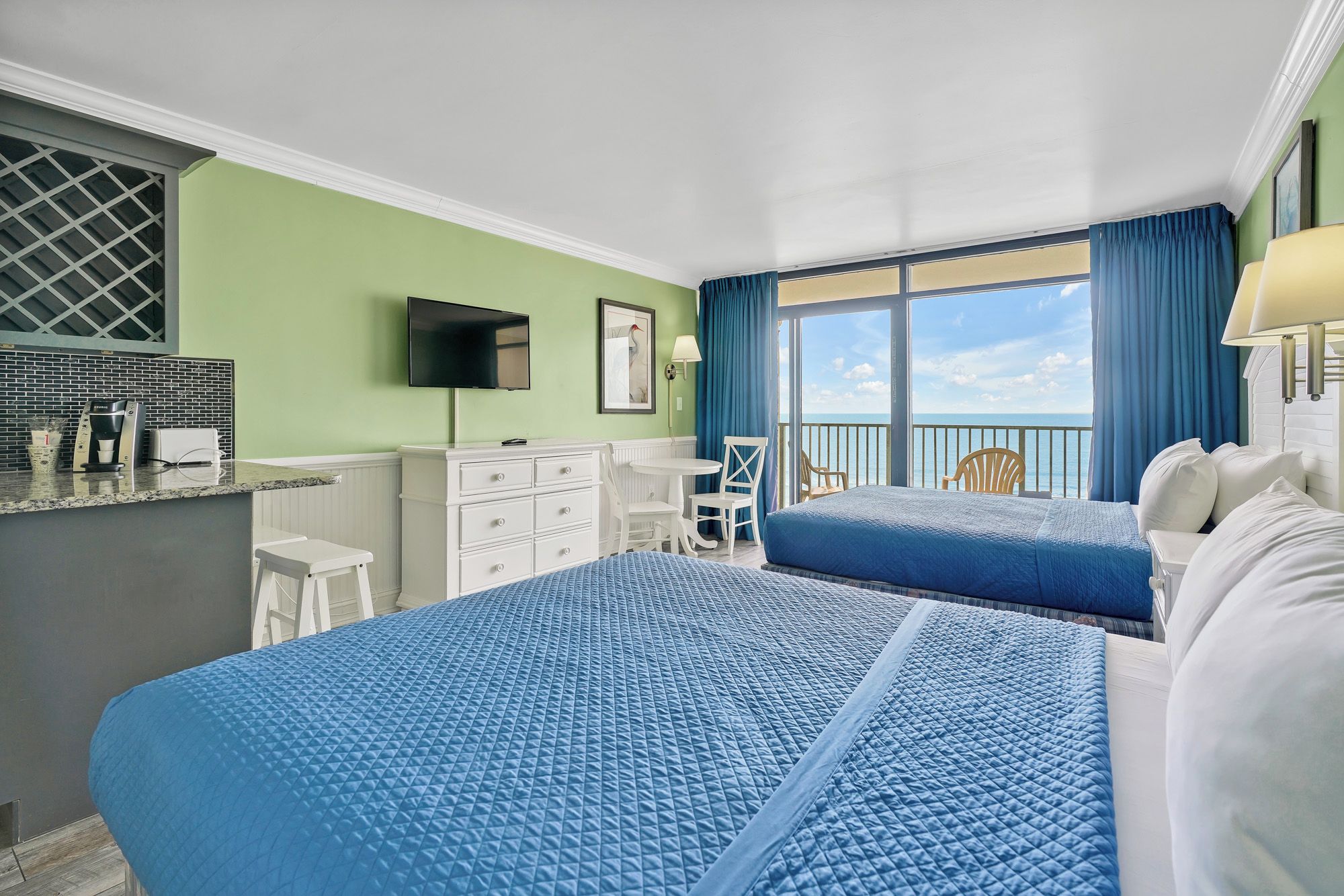 A hotel room with two beds, a flat-screen TV, a dining area, and a balcony with a view of the ocean.