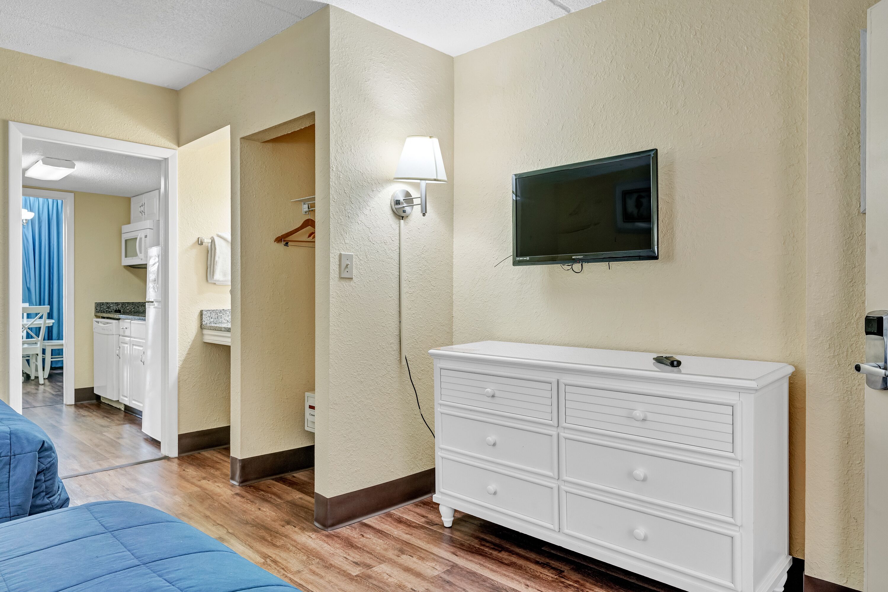 A hotel room with a wooden floor, a white dresser, a flat-screen TV mounted on the wall, and a hallway leading to a kitchen area.