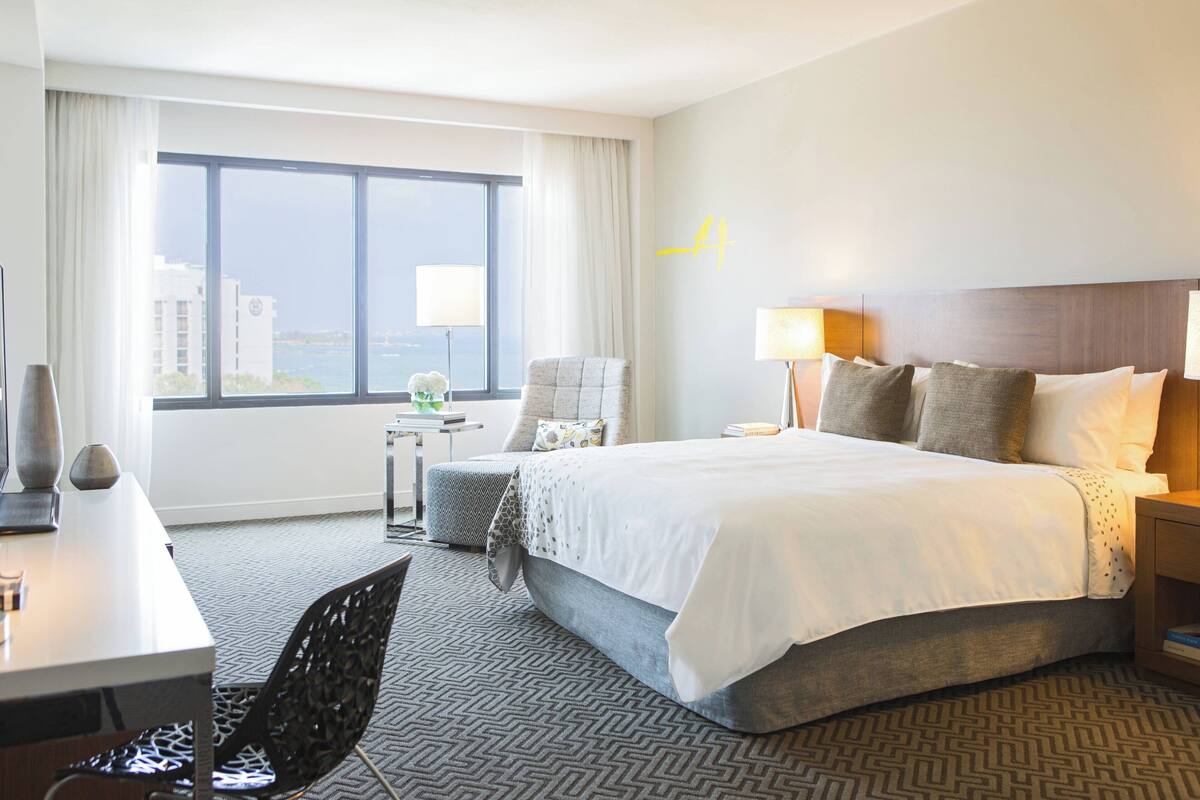 Une chambre d’hôtel avec un grand lit, un bureau, une chaise, une table de chevet, une lampe et une vue sur la ville par la fenêtre.
