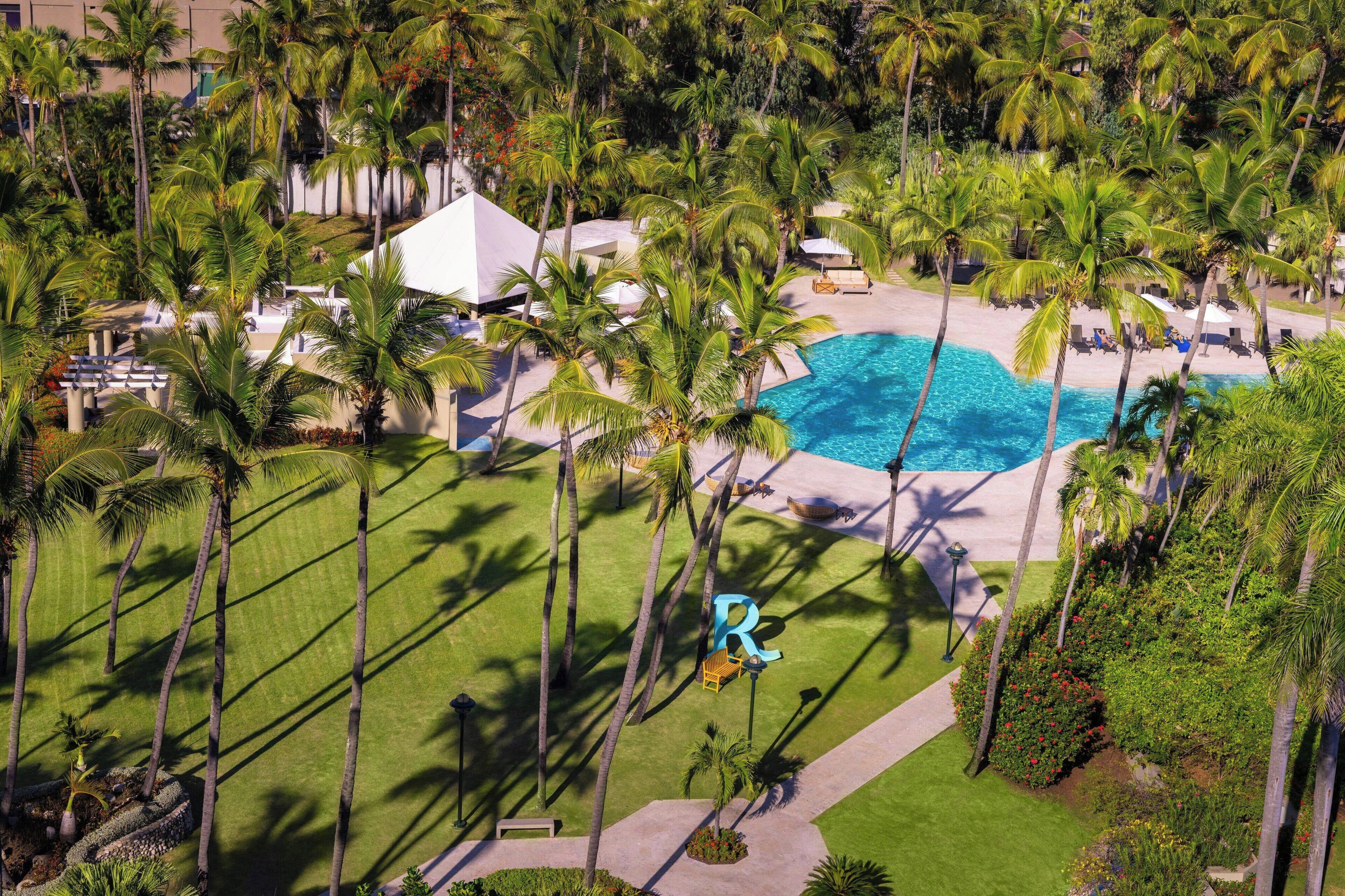 An aerial view of a resort with a swimming pool, palm trees, and a white building.