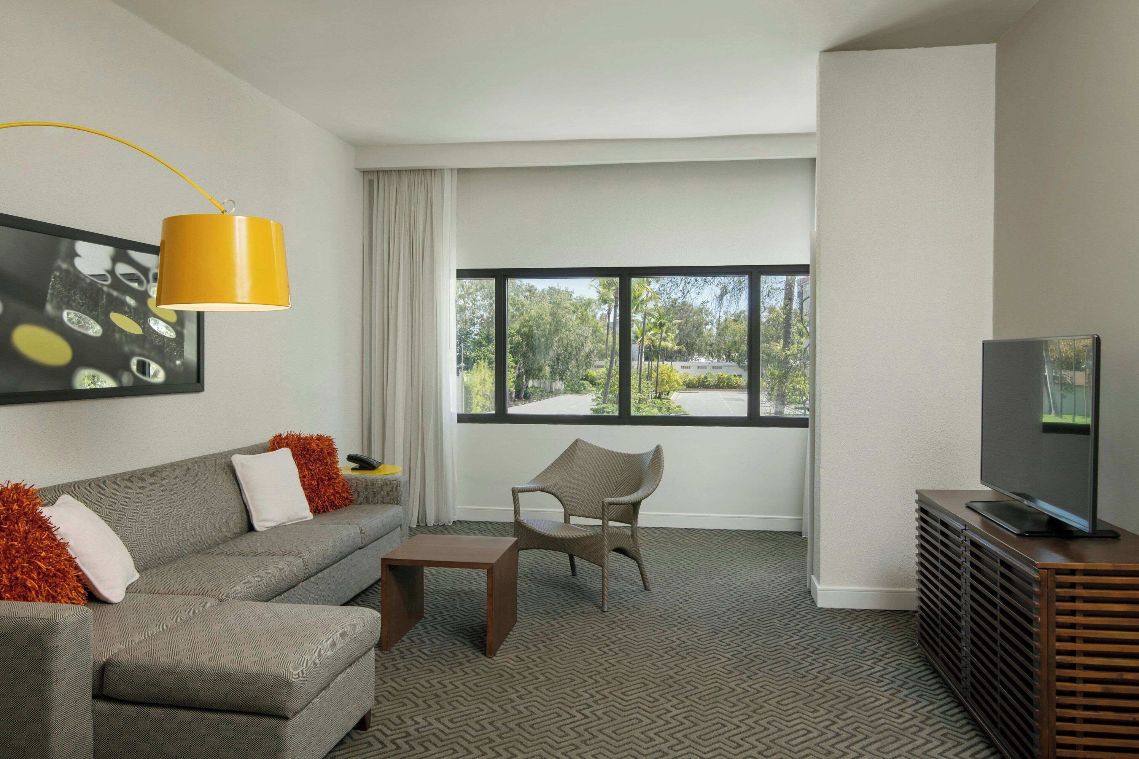 A modern living room with a sofa, armchair, coffee table, TV, and a large window with a view of greenery.