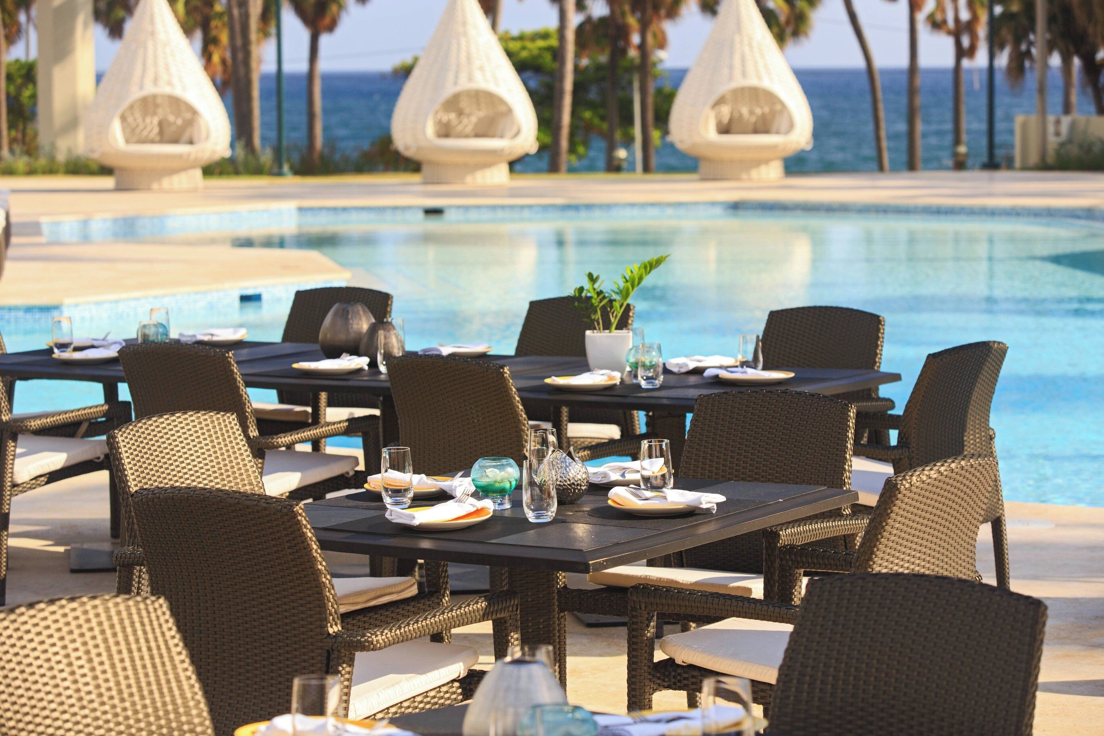 Un espace repas aménagé au bord de la piscine, avec des chaises et des tables en osier, une plante en pot et une vue sur l’océan.