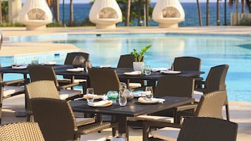 Un espace repas aménagé au bord de la piscine, avec des chaises et des tables en osier, une plante en pot et une vue sur l’océan.
