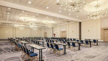 A large conference room with rows of tables and chairs, ceiling chandeliers, and a carpeted floor.