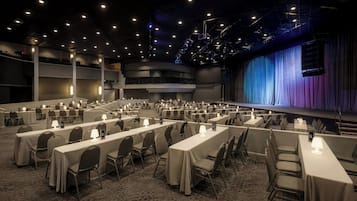 Un salón de conferencias con varias mesas y sillas, un escenario con cortinas y una zona de balcón.