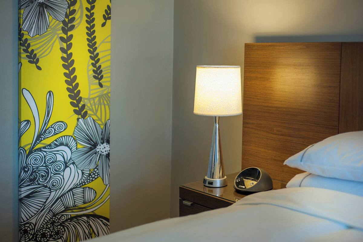Une chambre d’hôtel avec un lit, une table de chevet avec une lampe, une horloge et un mur orné d’un motif floral.