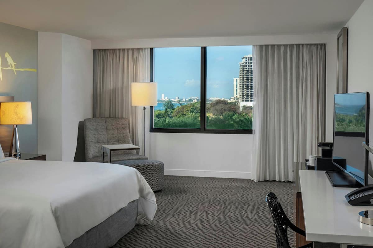Une chambre d’hôtel équipée d’un lit, d’un bureau, d’une chaise, d’une lampe, d’un téléviseur et offrant une vue sur le paysage urbain grâce à de grandes fenêtres.
