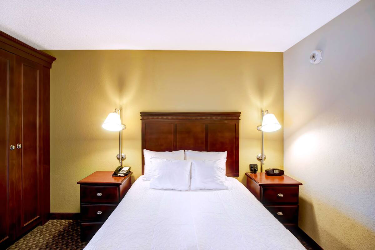 A hotel room with a large bed, two bedside tables, a wooden wardrobe, and two wall-mounted lamps.