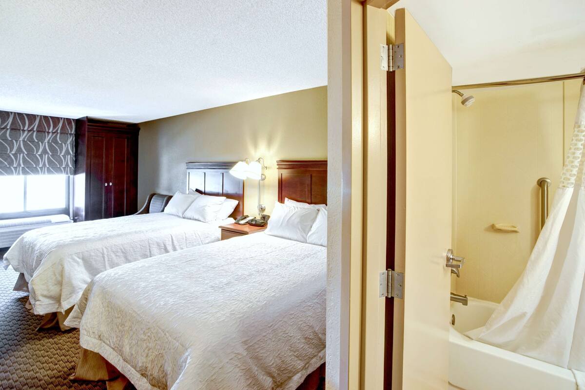 A hotel room with two beds, a nightstand, a lamp, and a shower area.