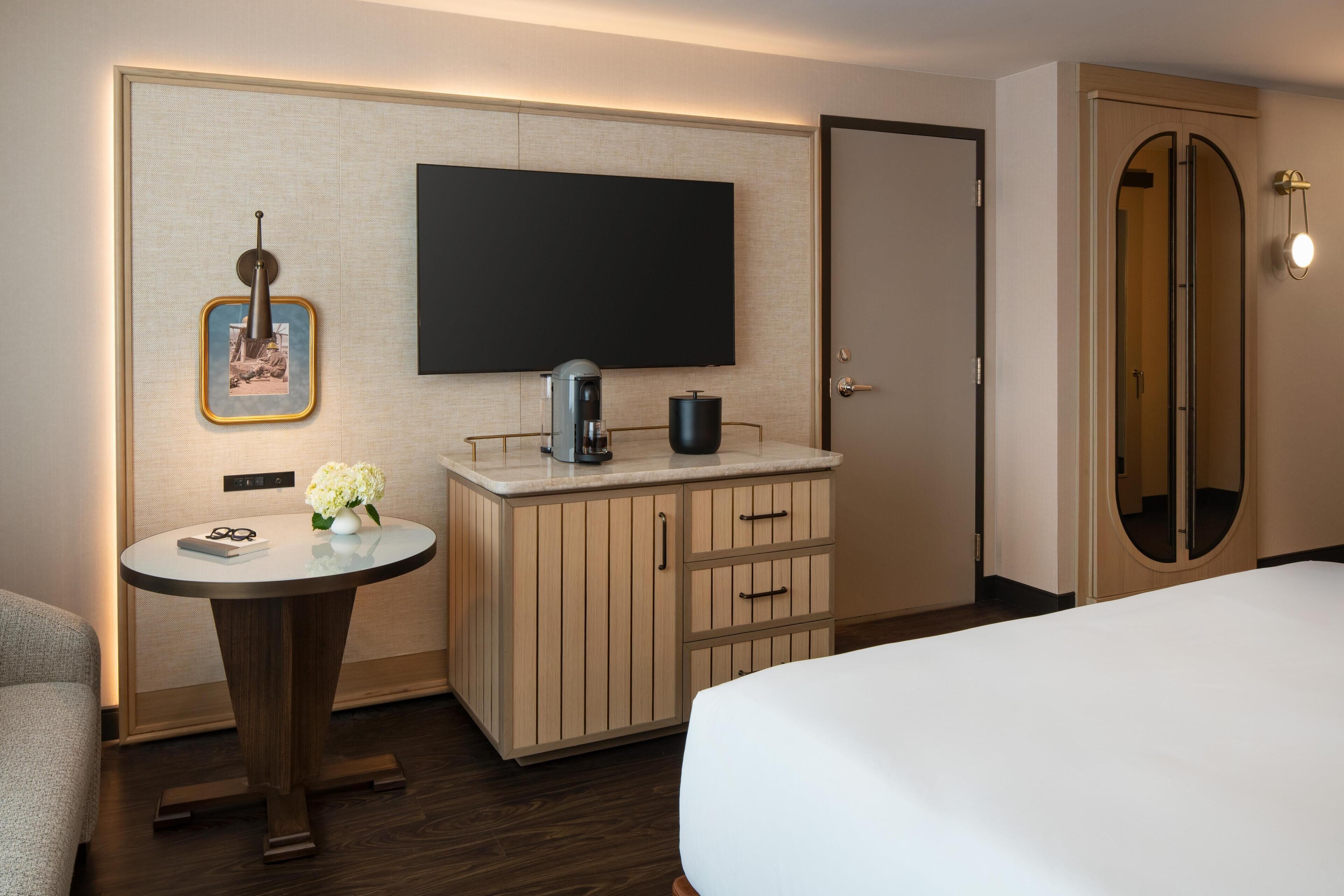A hotel room with a bed, a small round table with a coffee maker, a TV mounted on the wall, and a wardrobe with a mirror.