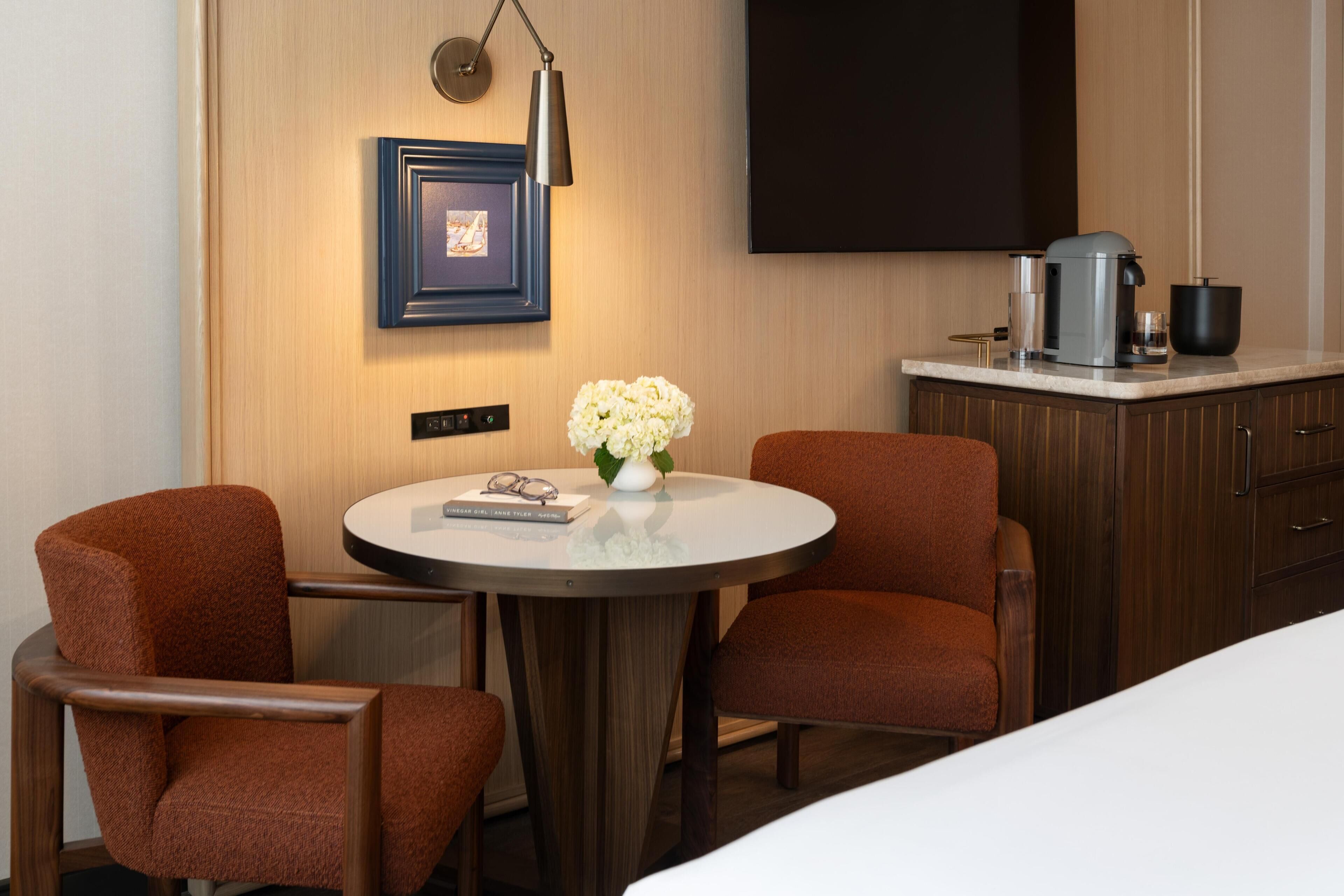 Una habitación de hotel con una mesa redonda, dos sillones, cafetera, televisor y un cuadro en la pared.
