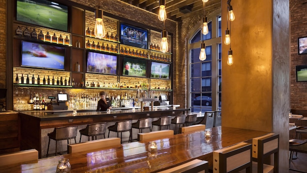 A bar area with multiple screens, a bar counter, seating, and a view through a large window.