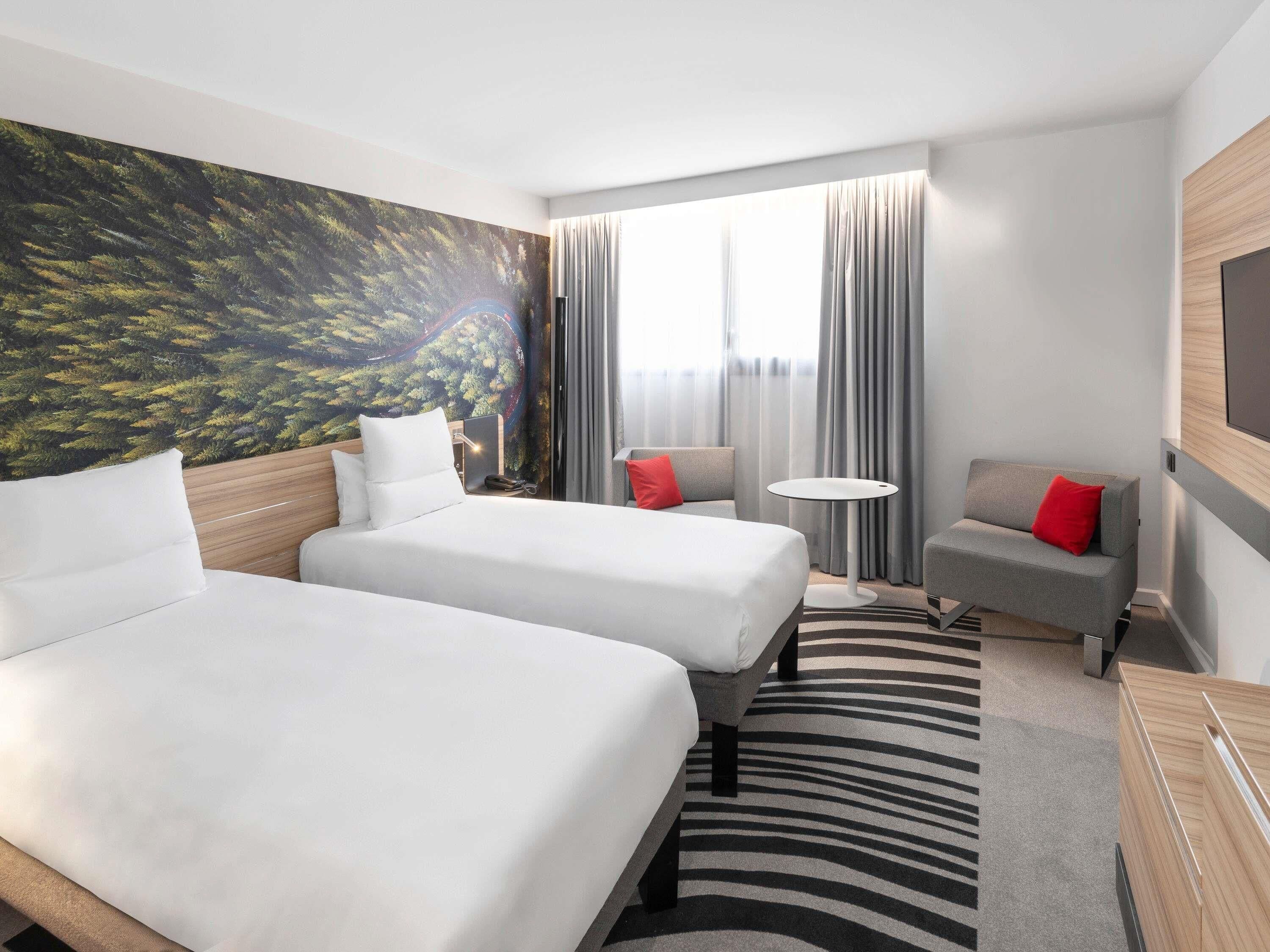 A hotel room with two beds, a small sofa, a round table, and a large window with curtains.