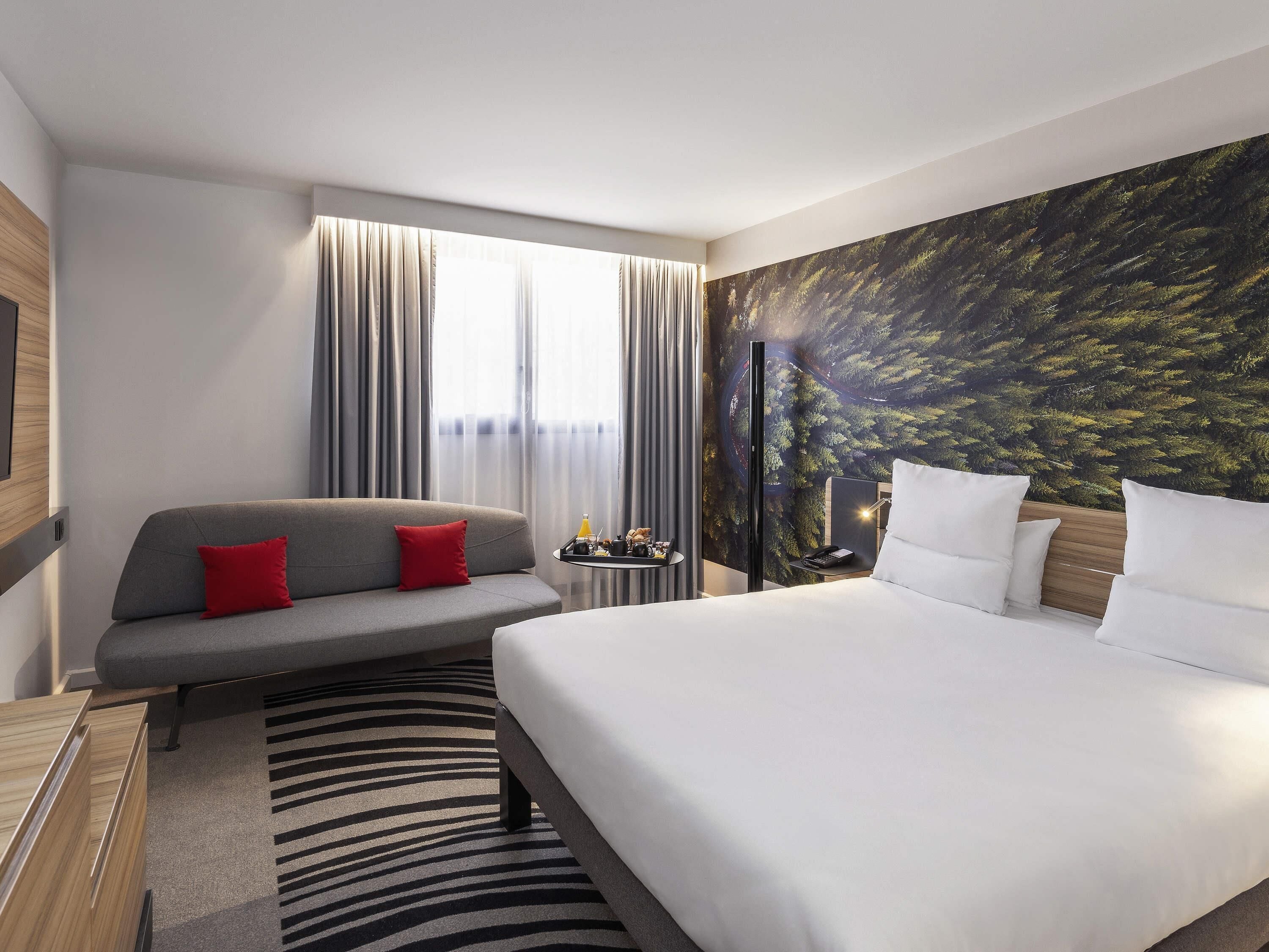 A hotel room with a bed, a sofa, a TV, a desk with a lamp, and a wall with a nature-themed mural.