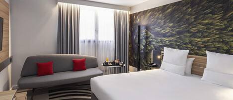 Una habitación de hotel con cama, sofá, televisor, escritorio con lámpara y un mural con motivos naturales.