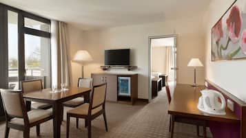 A hotel room with a dining table, chairs, a television, a minibar, and a painting on the wall.