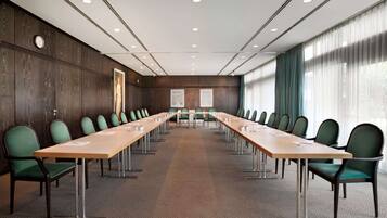 Un salón de conferencias con una larga mesa, sillas verdes, un reloj y una ventana con cortinas.