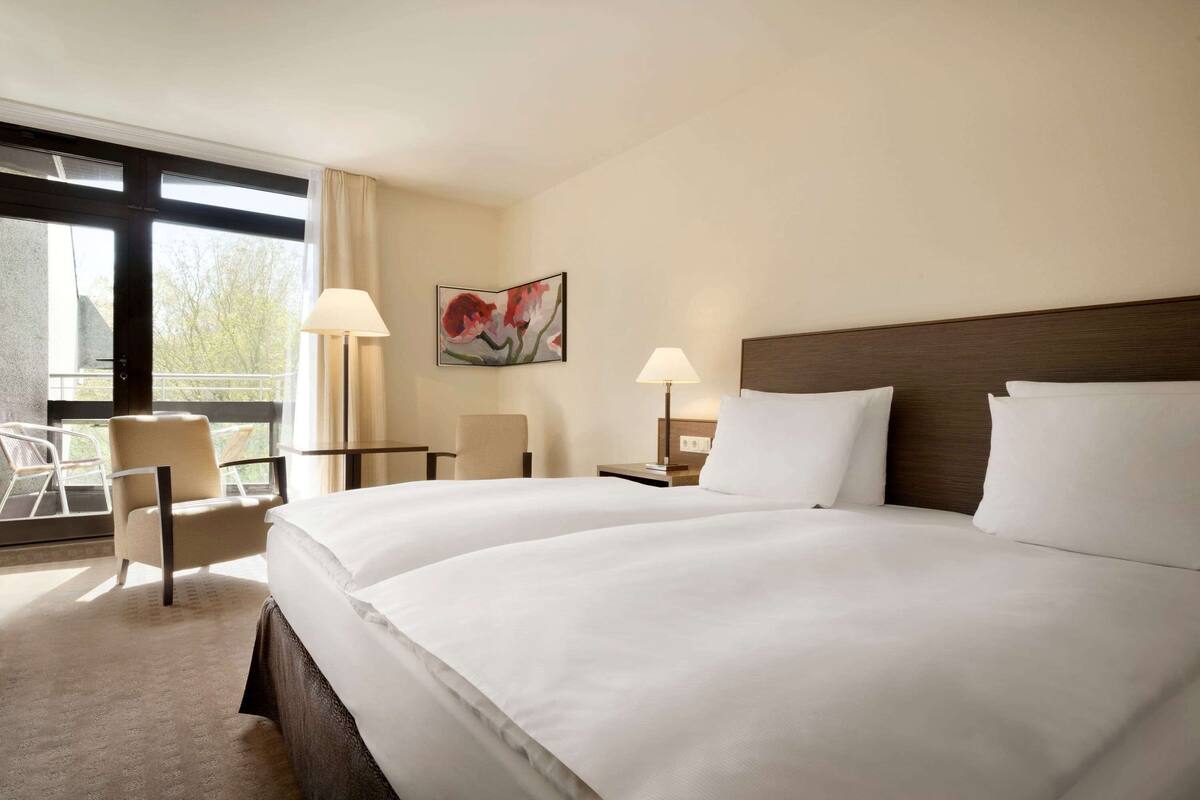 Une chambre d’hôtel avec un grand lit, deux tables de chevet avec des lampes, une chaise, un bureau et une fenêtre donnant sur des arbres.
