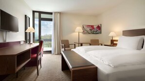 A hotel room with a large bed, a desk, a chair, a TV, a painting, and a window with curtains. - Ramada by Wyndham Nuernberg Parkhotel (Nuremberg)