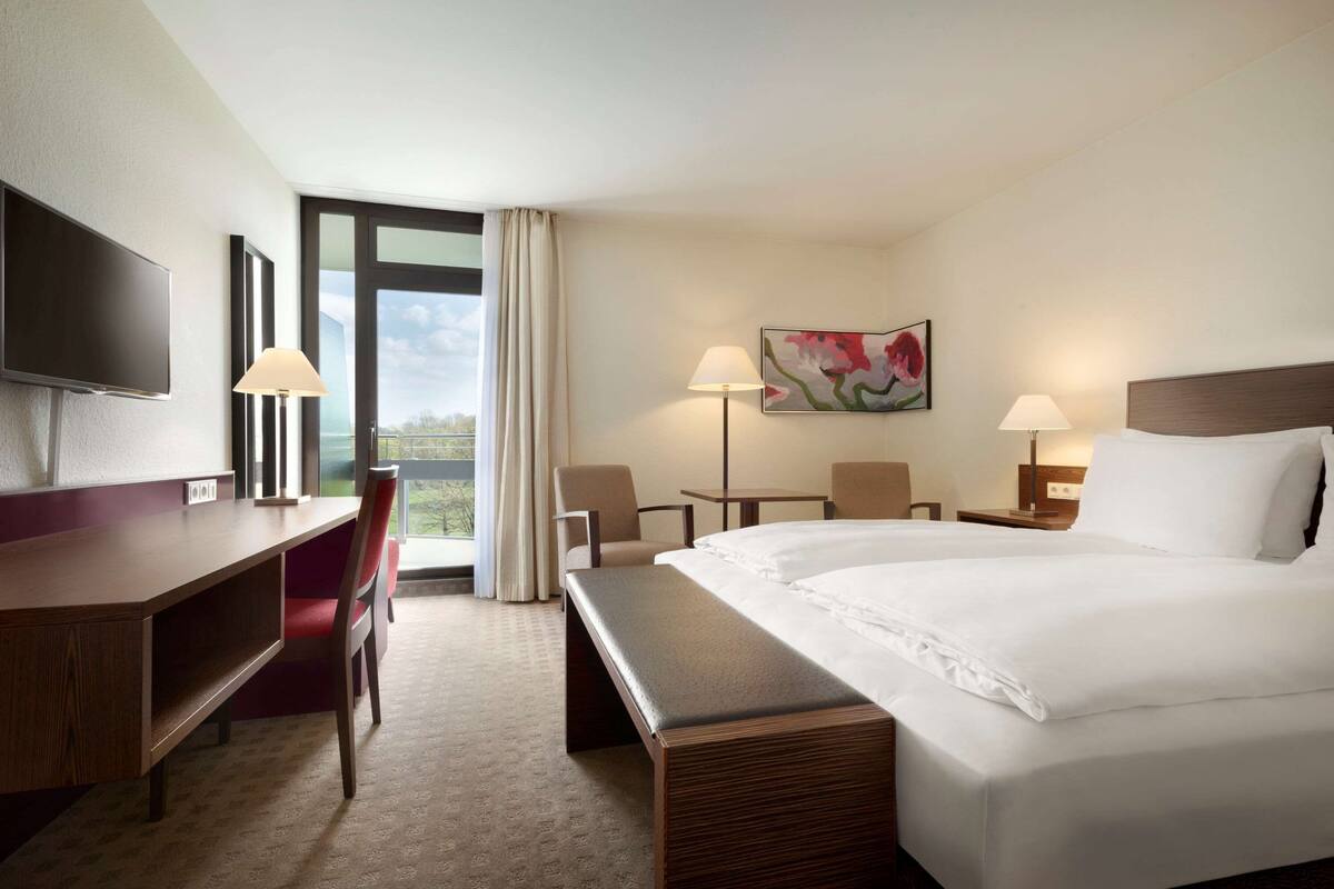 Une chambre d’hôtel avec un grand lit, un bureau, une chaise, une télévision, un tableau et une fenêtre avec des rideaux.