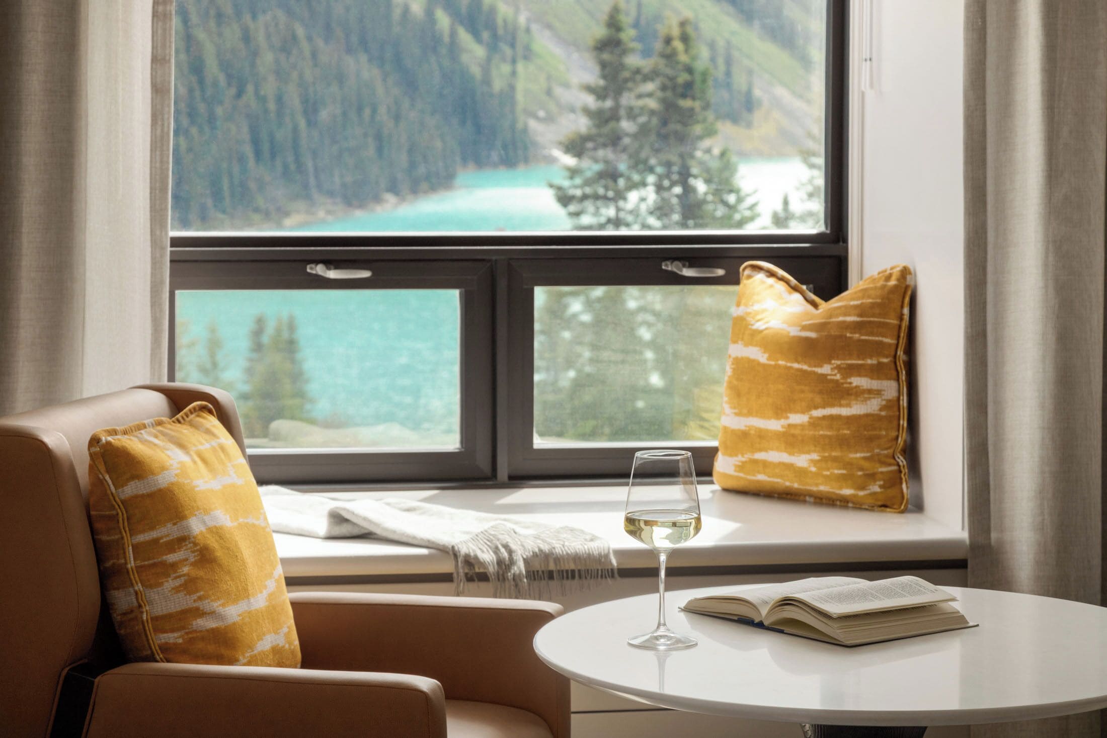 A room with a view of a lake and mountains, a window with two cushions, a glass of wine, and a book on a small table.