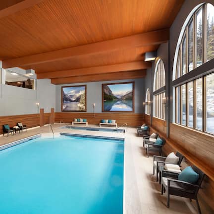 An indoor swimming pool with a wooden ceiling, seating area with chairs and cushions, and large windows with mountain views.