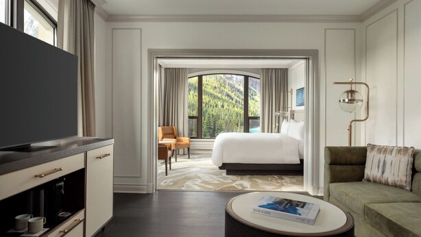 Ein Hotelzimmer mit einem großen Bett, einem Fernseher, einer Couch und einem Couchtisch. Durch das Fenster ist ein Blick auf einen Wald zu sehen.