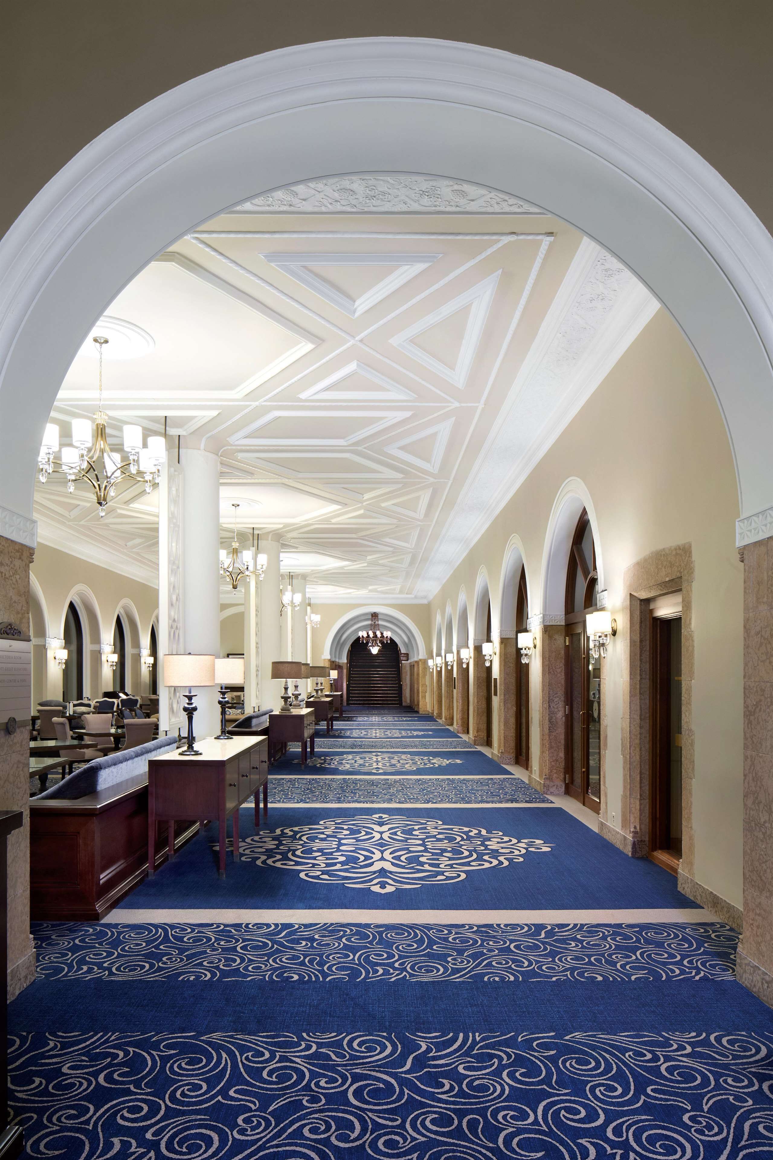 A hotel corridor with a blue patterned carpet, white columns, chandeliers, and arched doorways.