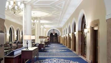 A hotel corridor with a blue patterned carpet, white columns, chandeliers, and arched doorways.
