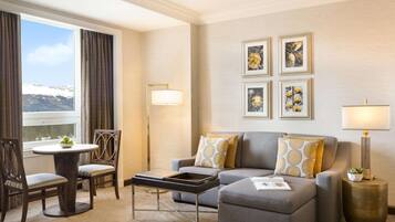 A modern hotel room with a grey sofa, a round dining table with chairs, a small side table, and a large window with curtains.
