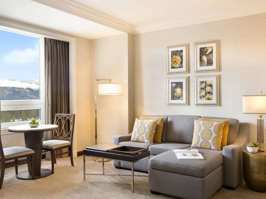 A modern hotel room with a grey sofa, a round dining table with chairs, a small side table, and a large window with curtains.