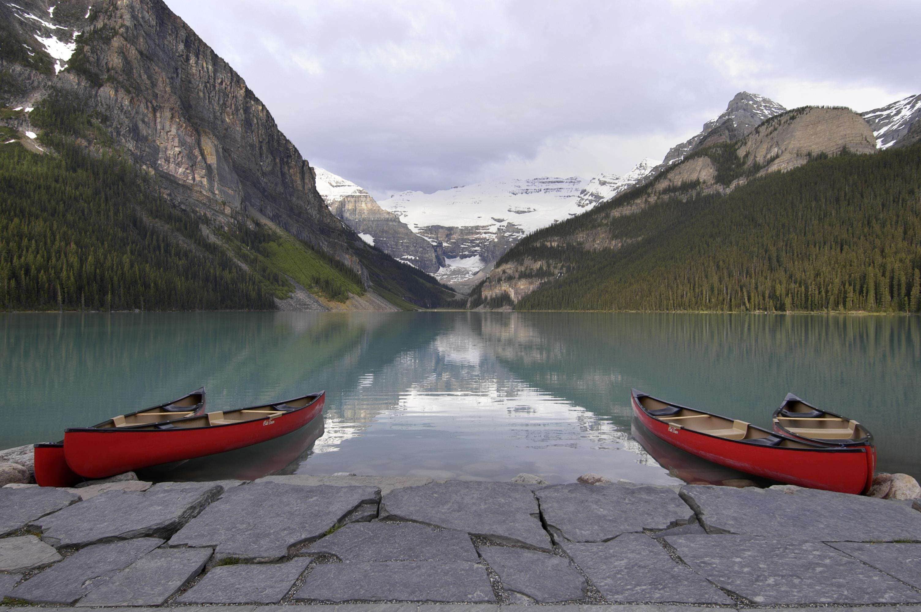 A calm lake with two red canoes on a stone dock, surrounded by forested mountains and snow-capped peaks.