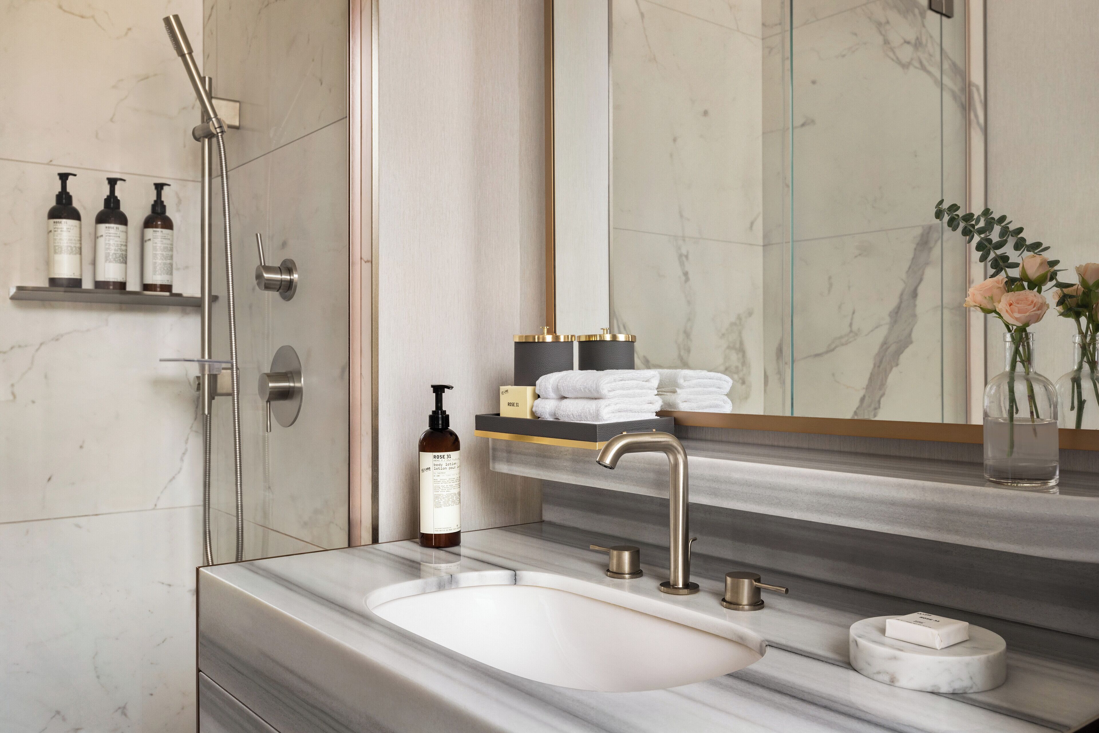Une salle de bain moderne avec un grand miroir, un lavabo en marbre et une douche. On trouve des articles de toilette sur une étagère et un vase avec des fleurs sur le comptoir.