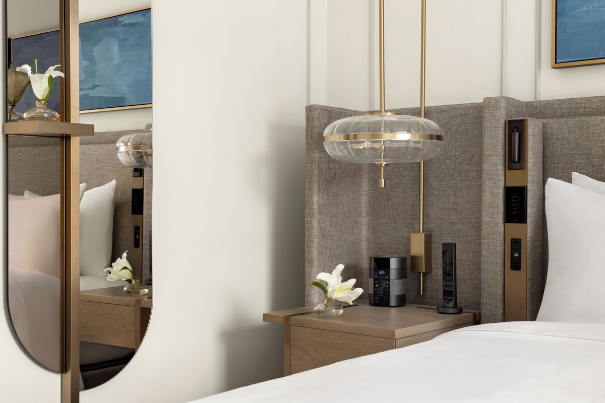 Une chambre d’hôtel moderne avec un grand lit, une table de chevet ornée d’un vase de fleurs, un panneau de commande fixé au mur et un grand miroir.