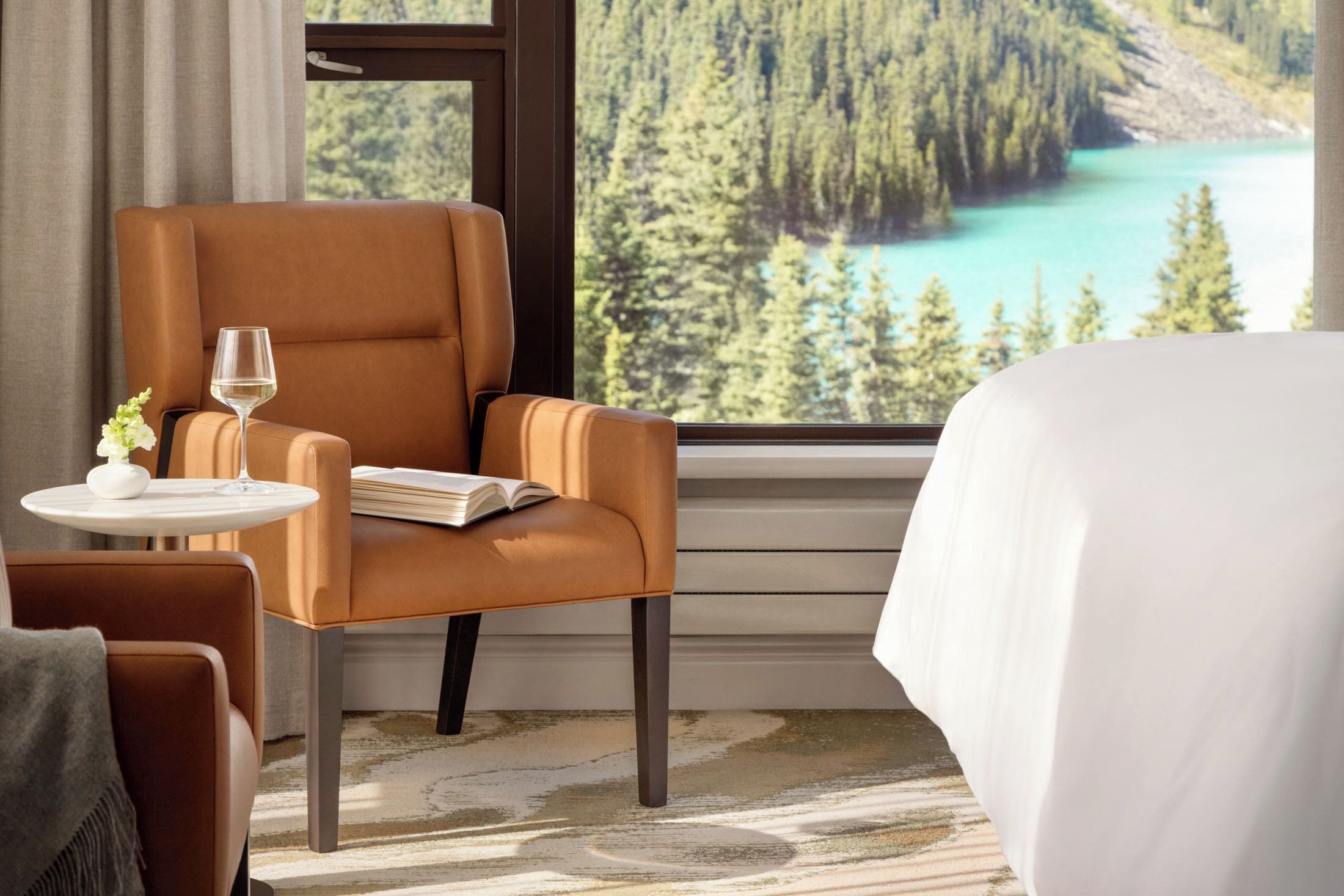 A hotel room with a large window offering a view of a forest and a lake. There is a brown armchair with a book and a glass of water on a small table beside it.