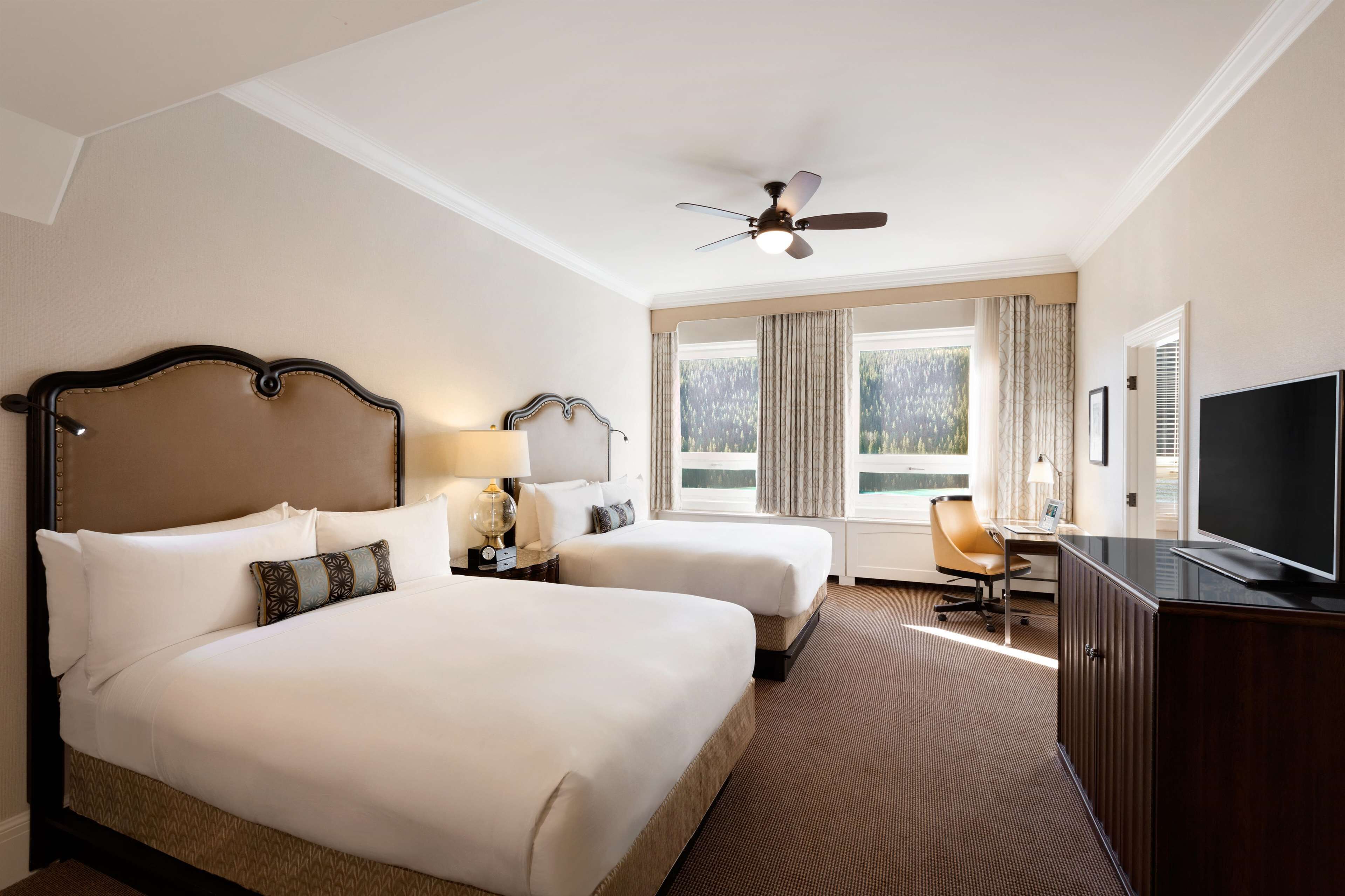 A hotel room with two beds, a desk with a television, a ceiling fan, and a view of the outdoors through large windows.