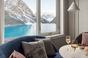 A room with a view of a lake and mountains, a blue sofa, two glasses of wine, and a table with a bowl of fruit.